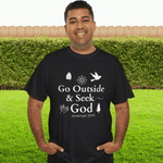 Man wearing a black t-shirt with 'Go Outside & Seek God' text, standing in a grassy area with a hedge.